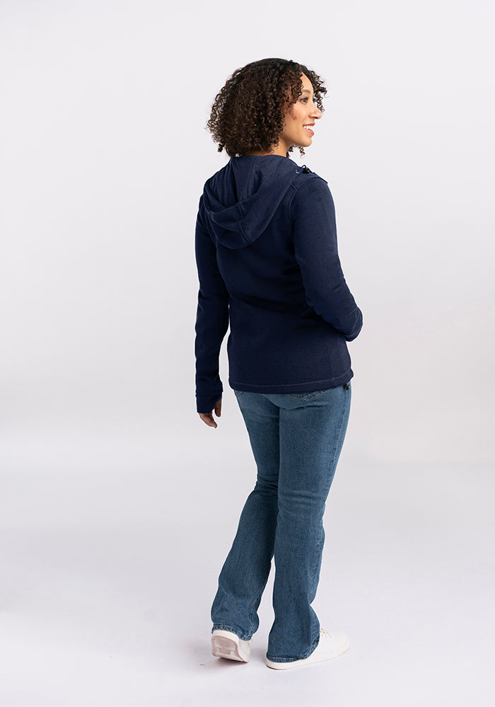 A person with curly hair stands with their back slightly turned to the side, smiling. They are wearing a deep navy Cubby Hooded Sweatshirt by Woolx, blue jeans, and white shoes against a plain white background.