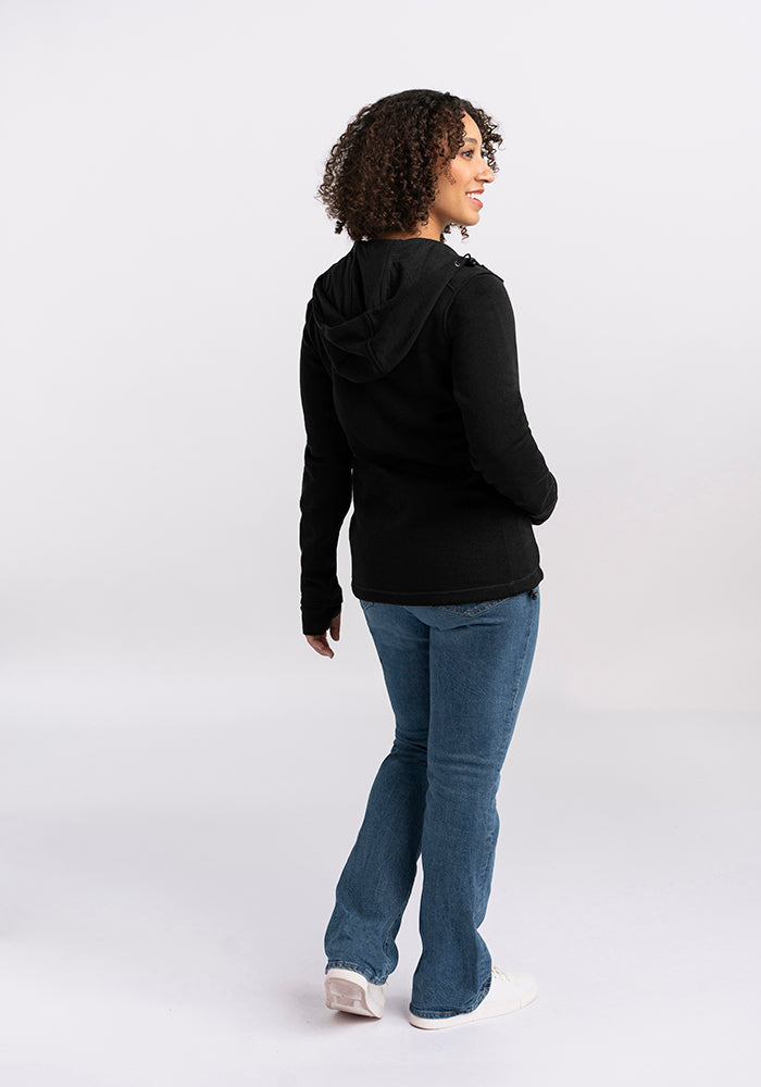 A person with curly hair wearing a Woolx Cubby Hooded Sweatshirt in black and blue jeans is standing against a plain white background. The individual is facing slightly away from the camera, gazing to the side, with one hand resting in their pocket and the other hanging naturally by their side.