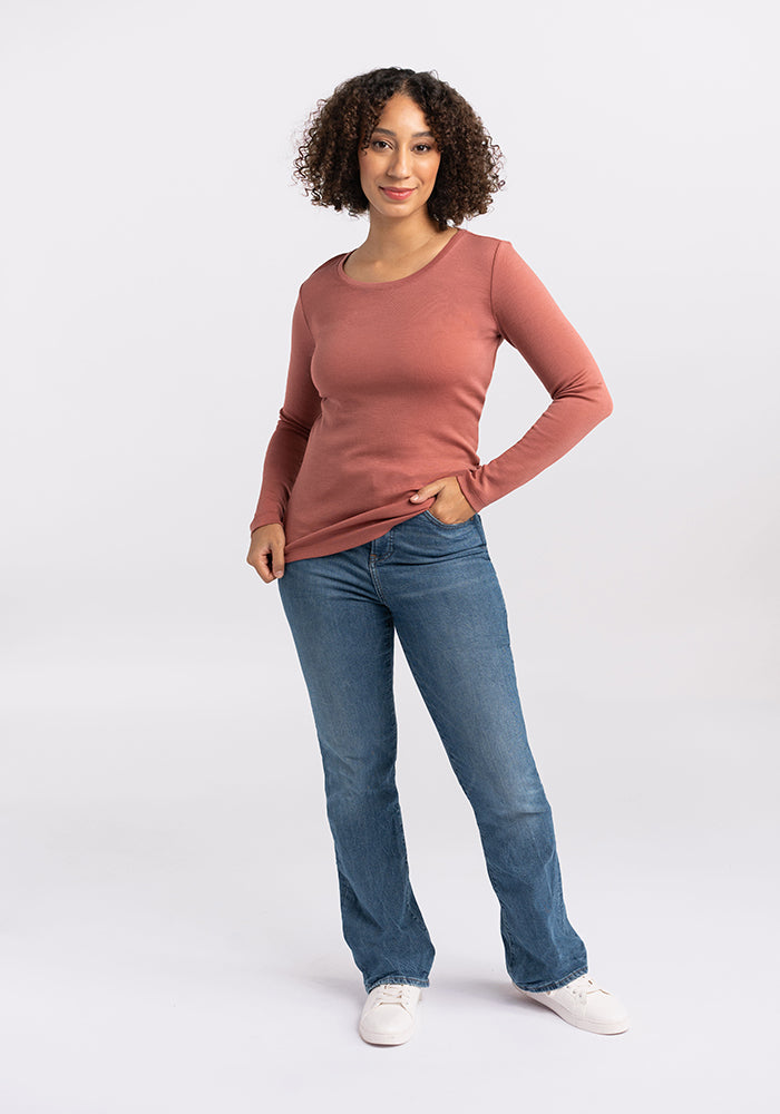 A person with short curly hair is standing against a plain white background, exuding business casual charm. They are wearing the fitted, long-sleeve Kenzie Scoop Neck top from Woolx in dusty pink, paired with blue jeans and white sneakers. One hand is resting on their hip as they flash a slight smile.