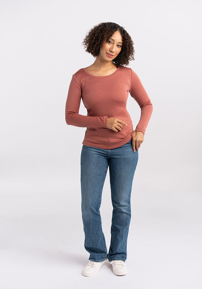 A person with curly hair stands against a white background, wearing a long-sleeved Kenzie Scoop Neck Merino wool top by Woolx in rust color, blue jeans, and white sneakers. They have one hand on their hip and the other resting by their side, looking directly at the camera with a neutral expression.