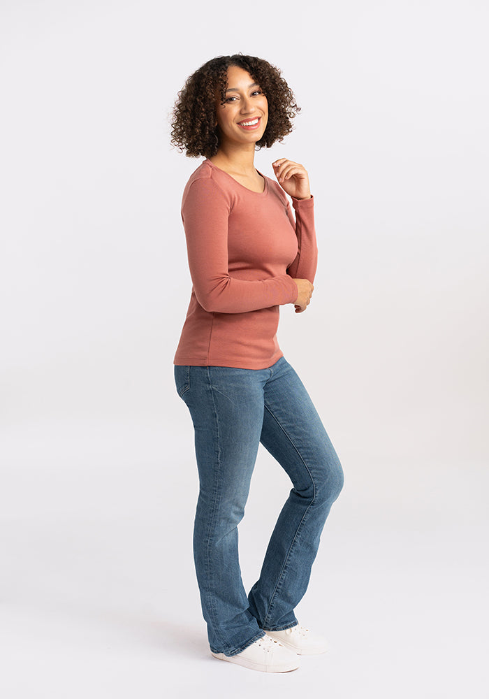 A person with curly hair, wearing a long-sleeve pink Kenzie Scoop Neck by Woolx, blue jeans, and white sneakers, stands with a smile. The individual is slightly turned to the side, with the left hand at the chin and right arm resting along the side. This business casual look fits perfectly for 3-seasons.