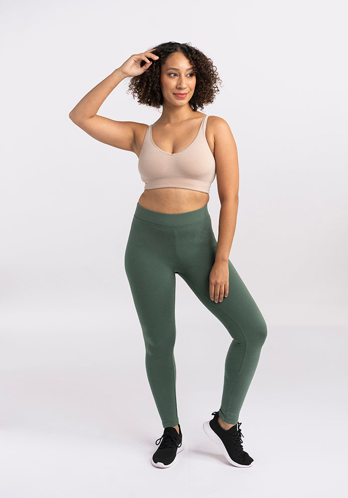 A woman with curly hair stands confidently with one hand resting on her head. She is wearing a beige sports bra, stylish green Stella Leggings by Woolx, and black athletic shoes against a plain white background.