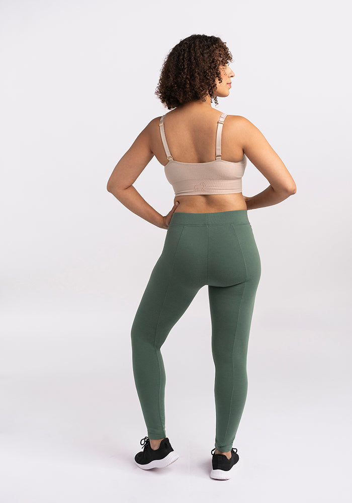 A person with curly hair stands facing away, showing their back. They are wearing a beige sports bra and green Stella Leggings by Woolx, paired with black sneakers—perfect for any winter wardrobe. The background is plain and white.