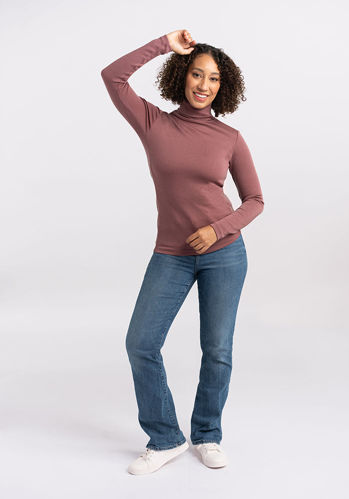 A person with curly hair stands confidently, smiling and posing with one hand on their head. They are wearing the Peyton Turtleneck from Woolx, a long-sleeve, high-neck maroon top made of merino wool, along with blue jeans and white sneakers. The background is a plain light gray, keeping the focus on the individual.