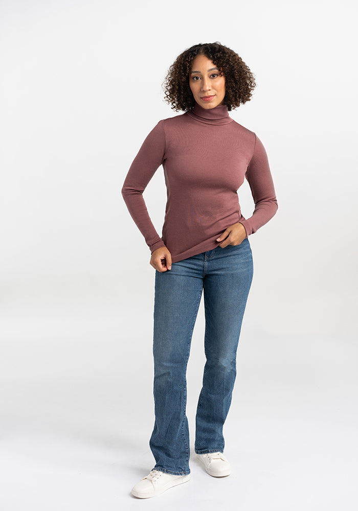 A person with curly hair is standing against a plain white background. They are wearing a long-sleeve, mauve Peyton Turtleneck from Woolx, blue jeans, and white sneakers. They have one hand resting on their hip and the other lightly holding the hem of their sweater, showcasing its warmth and comfort.