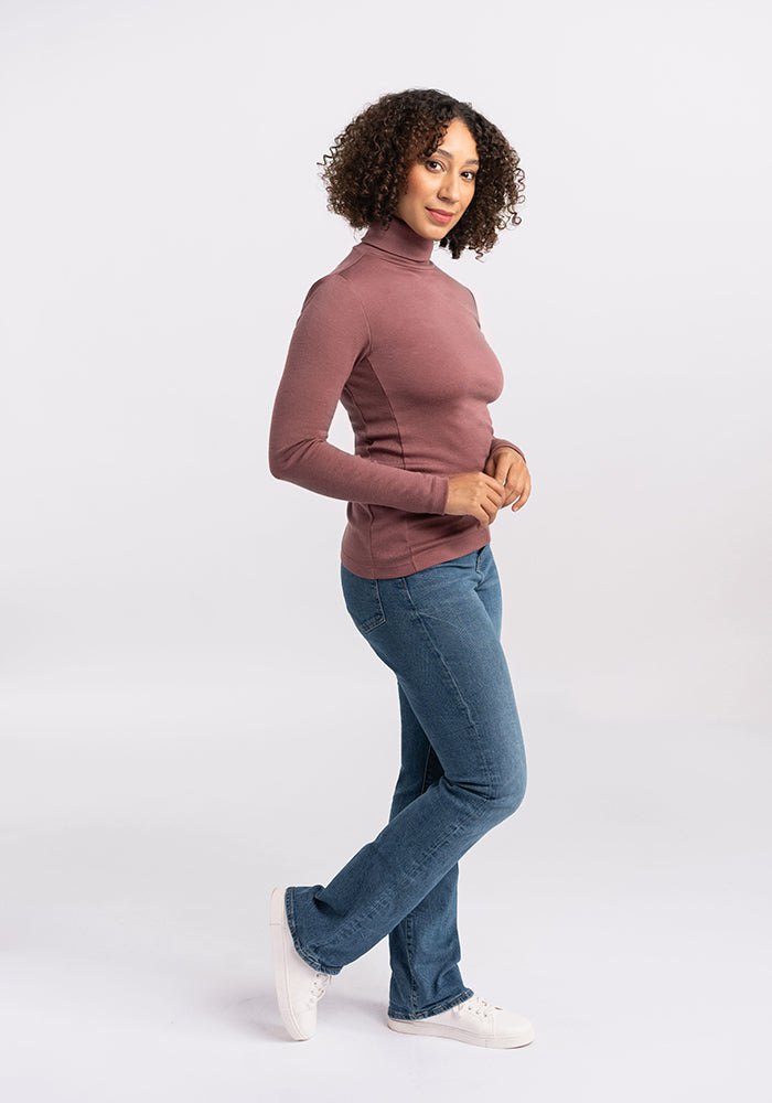 A person with curly hair stands against a plain background, wearing a long-sleeved, mauve Peyton Turtleneck from Woolx, paired with blue jeans and white sneakers. They are posed in a side profile with their hands lightly clasped in front.