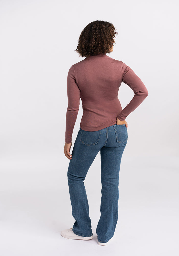 A person with curly hair is standing with their back to the camera, wearing a long-sleeved Peyton Turtleneck in dusty rose from Woolx and blue jeans. One hand rests on their hip while the other arm hangs by their side. The background is a plain, white studio setting.