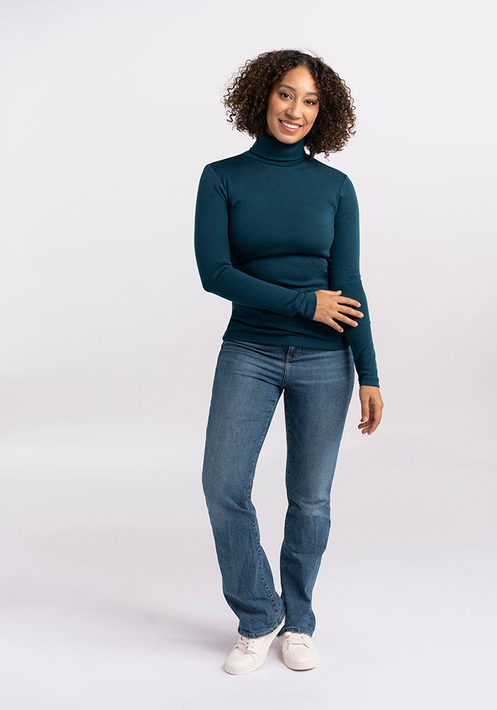 A person with curly hair is standing and smiling at the camera. They are wearing a dark teal Woolx Peyton Turtleneck made of midweight Merino wool, blue jeans, and white sneakers. Their right arm is bent at the elbow, with their left hand resting on their right forearm. The background is plain white.