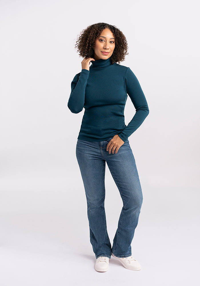 A woman with curly hair poses against a plain background, wearing a Woolx Peyton Turtleneck in dark teal, paired with blue jeans and white sneakers. One hand is at her chin while the other hangs by her side. She has a relaxed and confident expression.