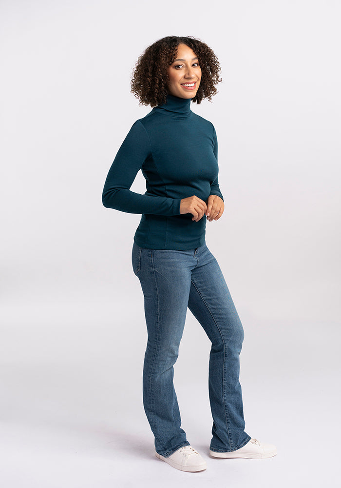 A person with curly hair is standing and smiling in a relaxed pose against a plain background. They are wearing the Peyton Turtleneck by Woolx, a dark teal midweight sweater made of Merino wool, paired with blue jeans and white sneakers.