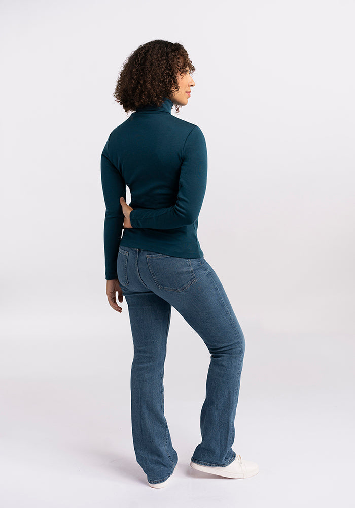 A person with curly hair stands facing slightly away from the camera, wearing a dark long-sleeve Peyton Turtleneck in Real Teal by Woolx and blue jeans. Their right hand is bent behind their back, and they are wearing white sneakers. The background is plain and light.