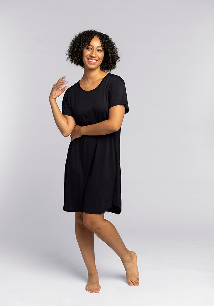 A person with curly hair stands barefoot against a plain background, wearing the Desi Nightgown in black from Woolx, which feels as comfy as temperature-regulating pajamas. They smile warmly with one hand raised near their shoulder, exuding an air of effortless comfort.