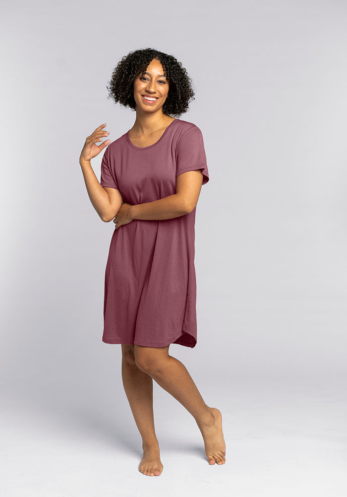 A person with curly hair smiles while standing barefoot against a plain grey background, elegantly dressed in the Desi Nightgown by Woolx, which is made of Merino wool and comes in a beautiful Wild Ginger color. Their right arm is bent, with the hand near their face, and their left arm rests by their side. 