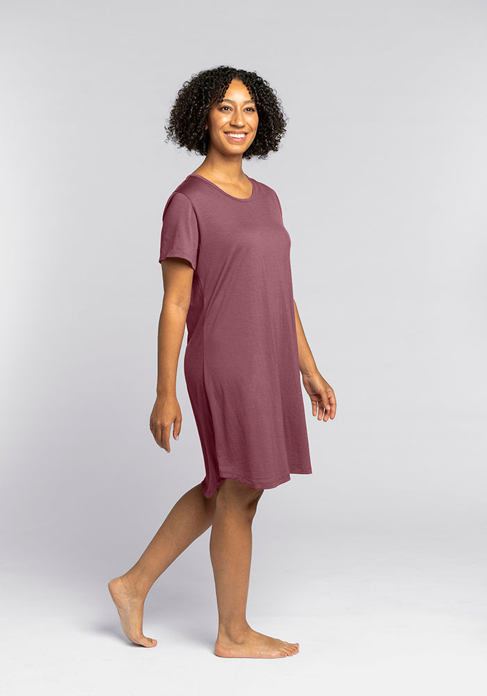 Wearing the Desi Nightgown - Wild Ginger from Woolx, a person with curly hair joyfully walks barefoot towards the camera. The casual, short-sleeved burgundy dress drapes comfortably on them as they smile against a plain light gray background.