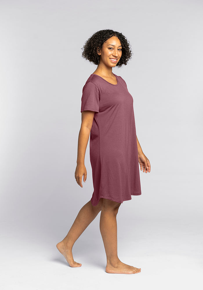 A person with curly hair is smiling and standing barefoot against a plain gray background. They are wearing the Desi Nightgown in Wild Ginger by Woolx, a comfortable loose, short-sleeve dress, and are slightly turned to the side with one foot stepped forward.