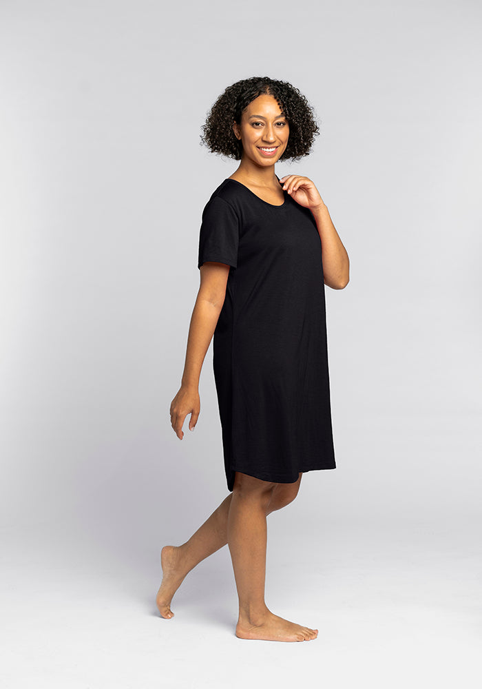 A person with curly hair smiles while standing barefoot in the uncomplicated elegance of the Desi Nightgown by Woolx. The black attire, akin to a cozy Merino wool dress, exudes comfort as they appear relaxed against a plain light gray background, perfectly capturing the essence of temperature-regulating sleepwear designed for blissful rest.