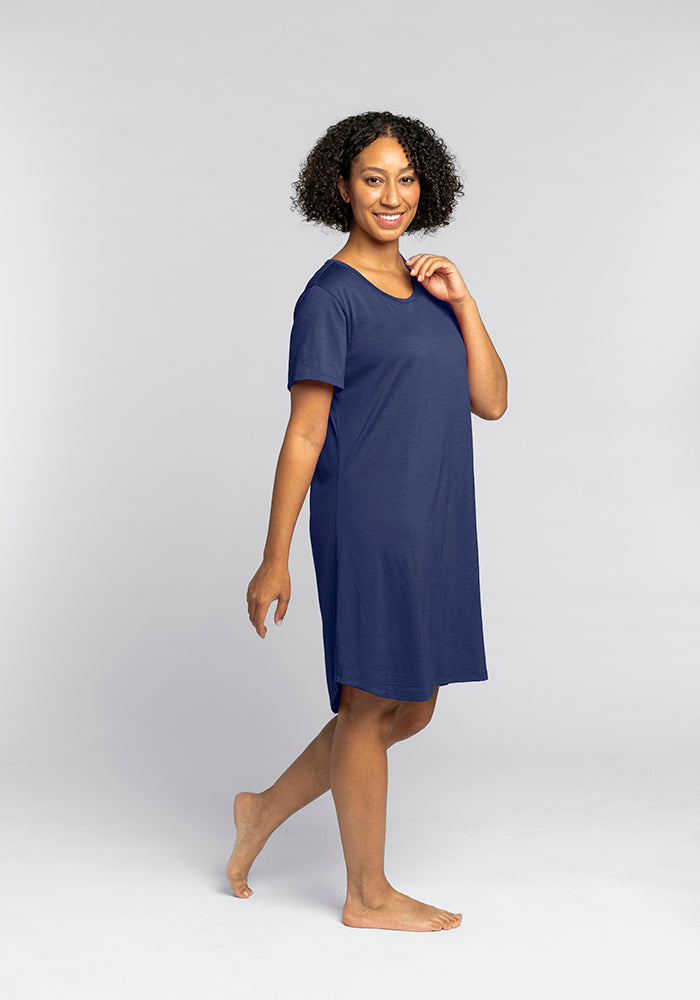 A person with curly hair smiles while wearing the Woolx "Desi Nightgown - Starry Night," a navy blue short-sleeved dress designed for temperature regulation. They are barefoot, standing against a plain light-gray background, and have one hand near their chin.