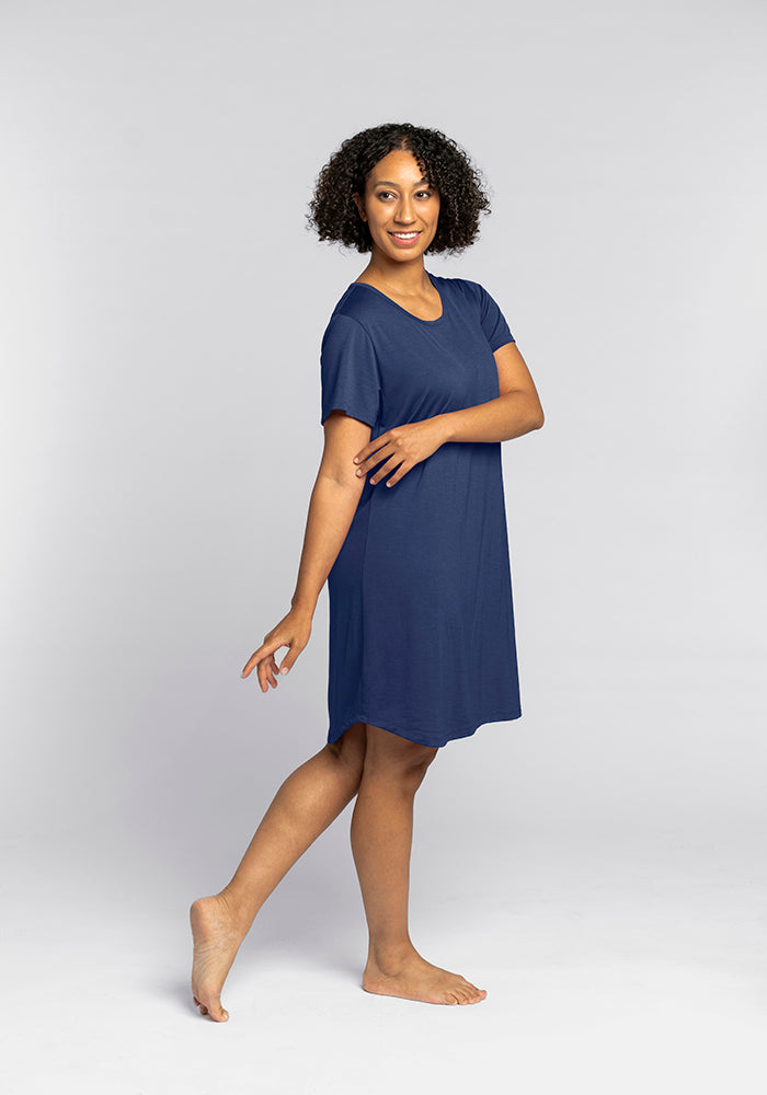 A person with curly hair is standing barefoot on a plain background, wearing the Desi Nightgown - Starry Night by Woolx. They are smiling and gracefully posing with one arm bent across their body and one foot slightly raised, exuding the cozy elegance of this temperature-regulating nightwear.