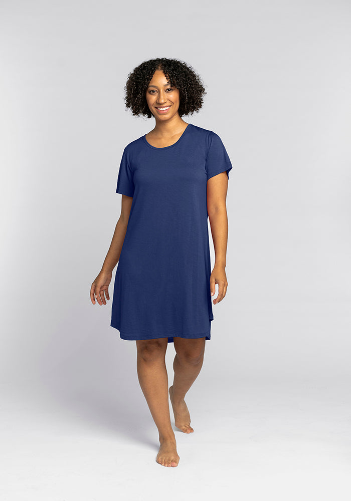 A person with curly hair is smiling and walking barefoot, wearing the cozy Desi Nightgown - Starry Night by Woolx, which resembles the comfort of merino wool sleepwear, set against a plain gray background. 