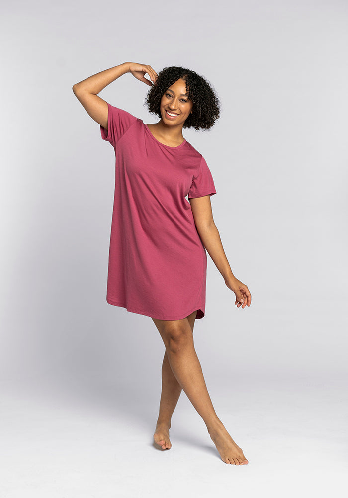 A person wearing Woolx's Desi Nightgown OLD, a short-sleeve, knee-length red dress crafted from comfortable merino wool, poses barefoot against a plain white background. They have curly hair and are smiling, with one arm raised and the other gently bent at their side.