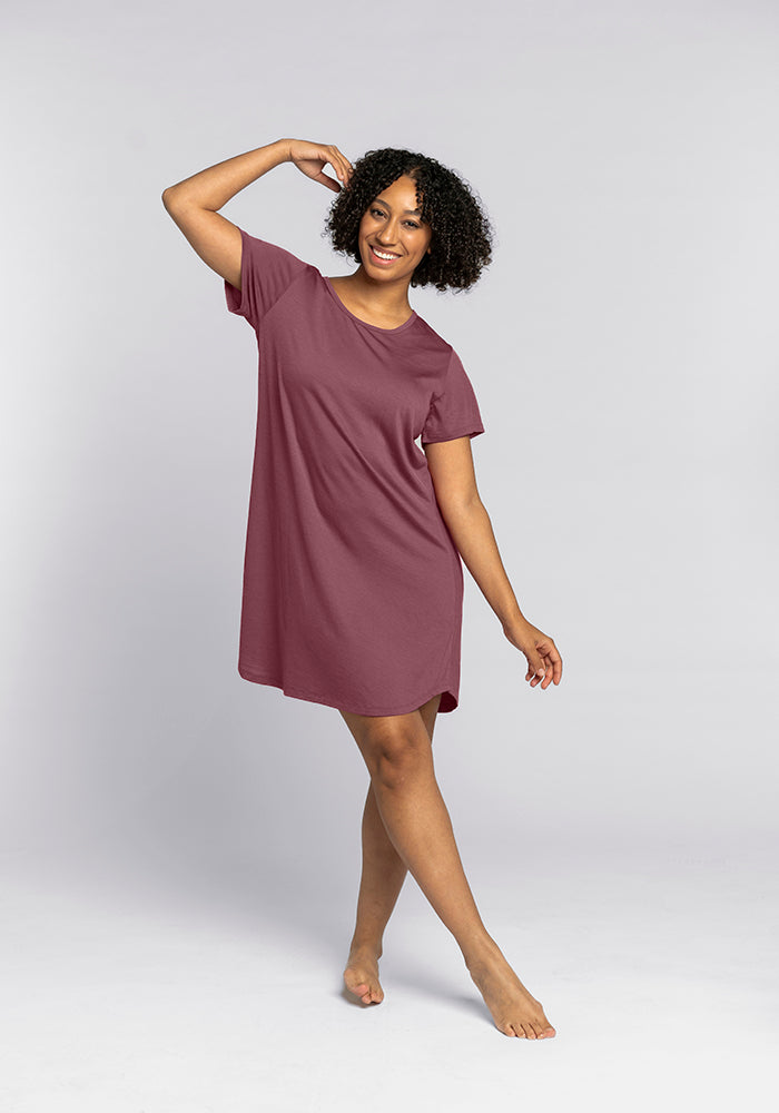 A woman with curly hair is smiling and wearing the Desi Nightgown in Wild Ginger from Woolx, a loose, short-sleeved dress that feels as cozy as comfort sleepwear. She poses confidently with one hand raised and the other relaxed by her side against a plain gray background.