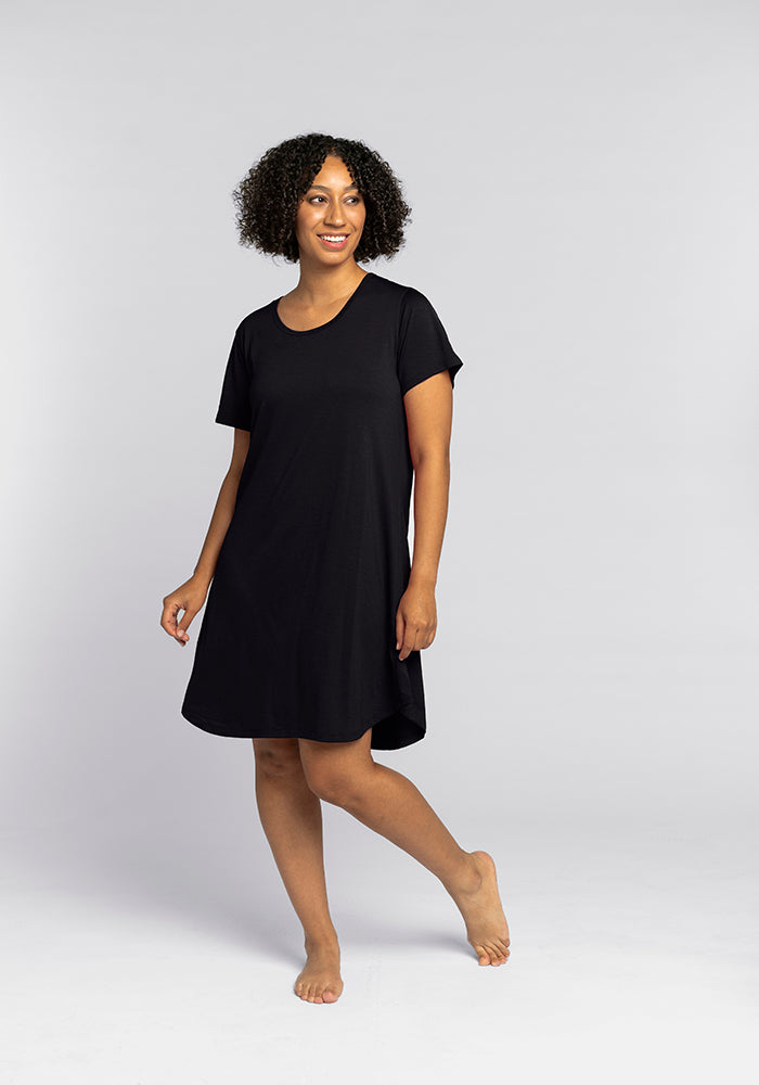 A person with curly hair is wearing the Desi Nightgown in black, a comfortable piece by Woolx that resembles temperature-regulating sleepwear. They are barefoot against a plain gray background, smiling and looking to the side.