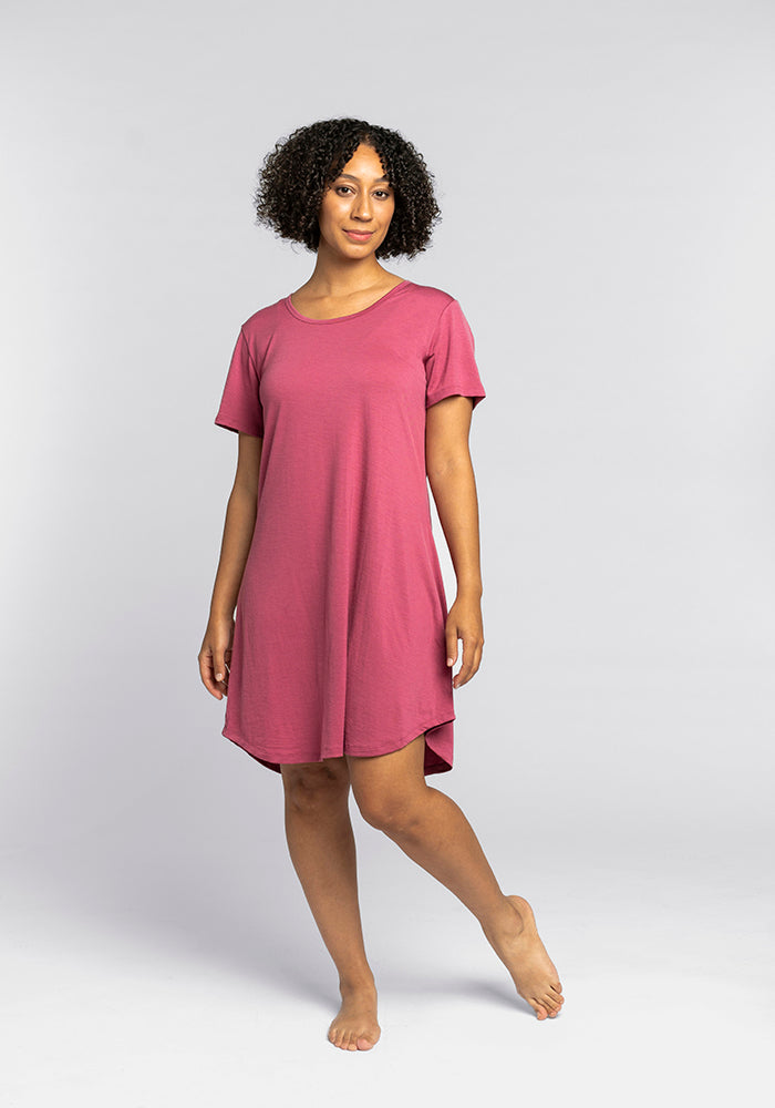 Wearing the Desi Nightgown OLD by Woolx, a person with curly hair is captured barefoot against a simple gray background. The short-sleeve pink dress resembles comfortable sleepwear, with one leg slightly bent as they softly smile at the camera, exuding cozy elegance.