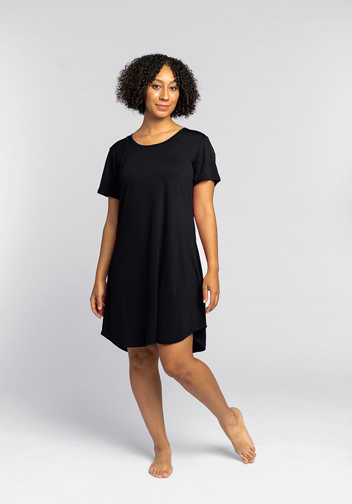 A person with curly hair poses confidently in the Desi Nightgown - Black by Woolx, their bare feet against a plain gray background, highlighting the elegance of temperature-regulating sleepwear. 