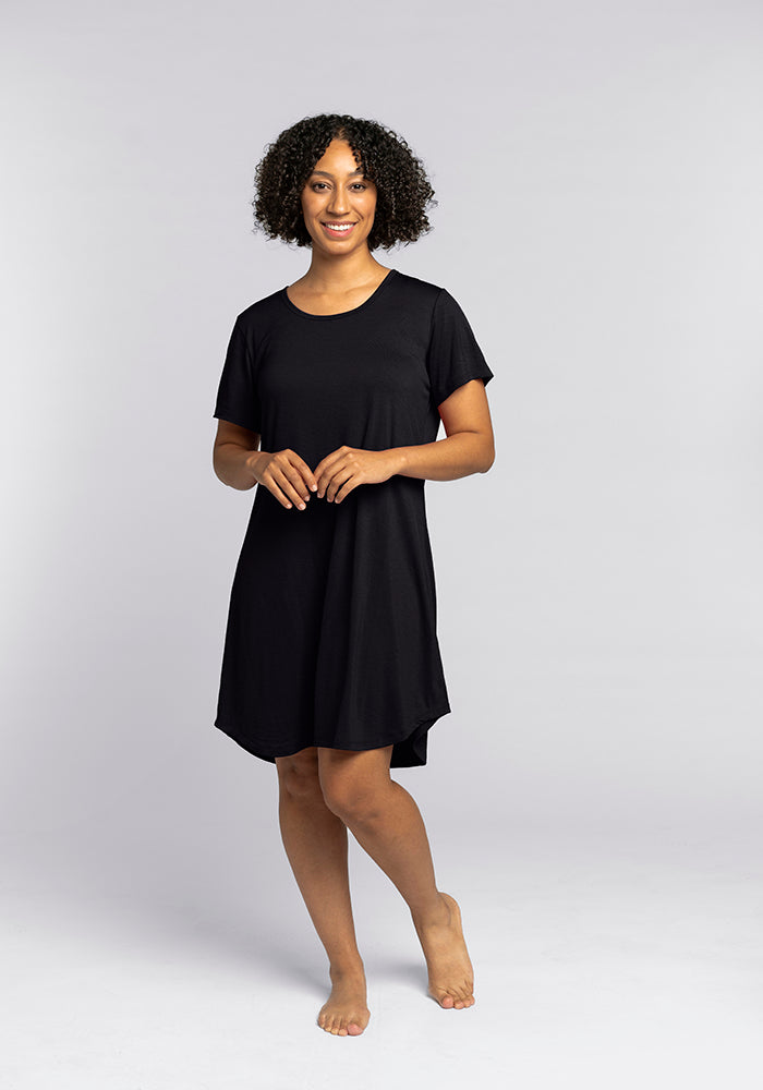 Against a gray backdrop, a person stands barefoot wearing the Desi Nightgown in black from Woolx. With curly hair and a friendly expression, they gently clasp their hands in front of them, embodying the comfort and ease of this Merino wool sleepwear design with its short sleeves.