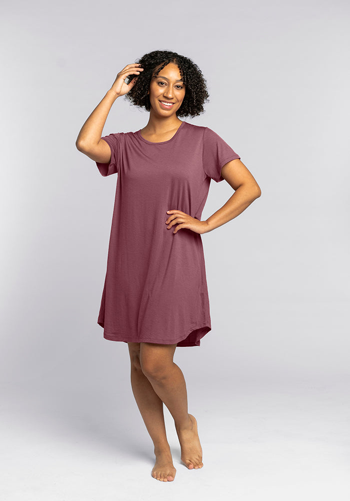 A person with curly hair smiles while posing in the simple, short-sleeved Desi Nightgown in Wild Ginger by Woolx. They stand barefoot against a plain gray background, with one hand touching their hair and the other on their hip, exuding the comfort of premium sleepwear.