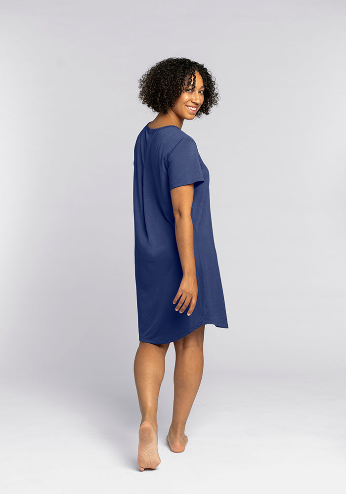 A person with curly hair is smiling and walking barefoot, wearing Woolx's Desi Nightgown - Starry Night, a short-sleeve blue dress known for its comfort. The plain light gray background enhances the serene vibe.