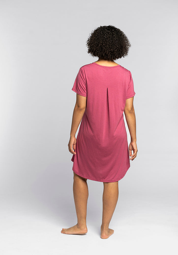 A person with curly hair stands barefoot against a gray background, showcasing the back of a short-sleeved, knee-length pink Desi Nightgown OLD by Woolx, designed for optimal temperature regulation.