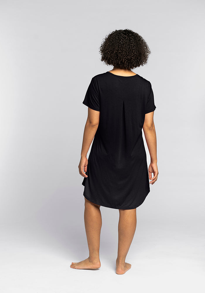 A person with curly hair is standing barefoot on a plain background, wearing the Woolx Desi Nightgown in black. Made from Merino wool, this short-sleeved dress offers temperature-regulating comfort as they face away from the camera.