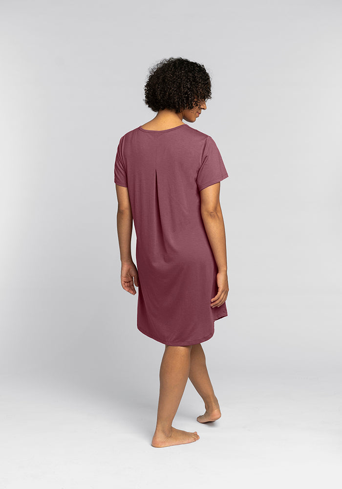 Rear view of a person with curly hair wearing the Woolx Desi Nightgown in Wild Ginger, a knee-length, short-sleeved dress made from merino wool. They are barefoot and standing on a white background, looking over their shoulder.