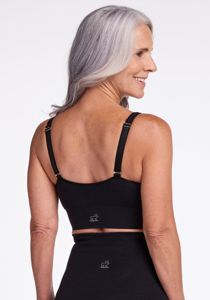 A woman with long gray hair is smiling and standing with her back to the camera, wearing a black Hayden Bra from Woolx, featuring adjustable straps. She is also wearing a matching black bottom. The plain white background emphasizes the comfort of this lounge essential.