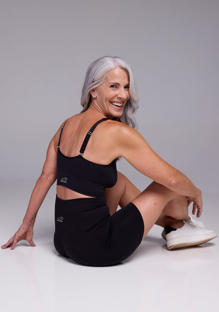 A smiling woman with long gray hair sits on the floor, turned slightly to the side. She is wearing a black sports outfit featuring the Woolx Hayden Bra with a cutout on the back and white sneakers. The background is a plain, light gray studio setting.