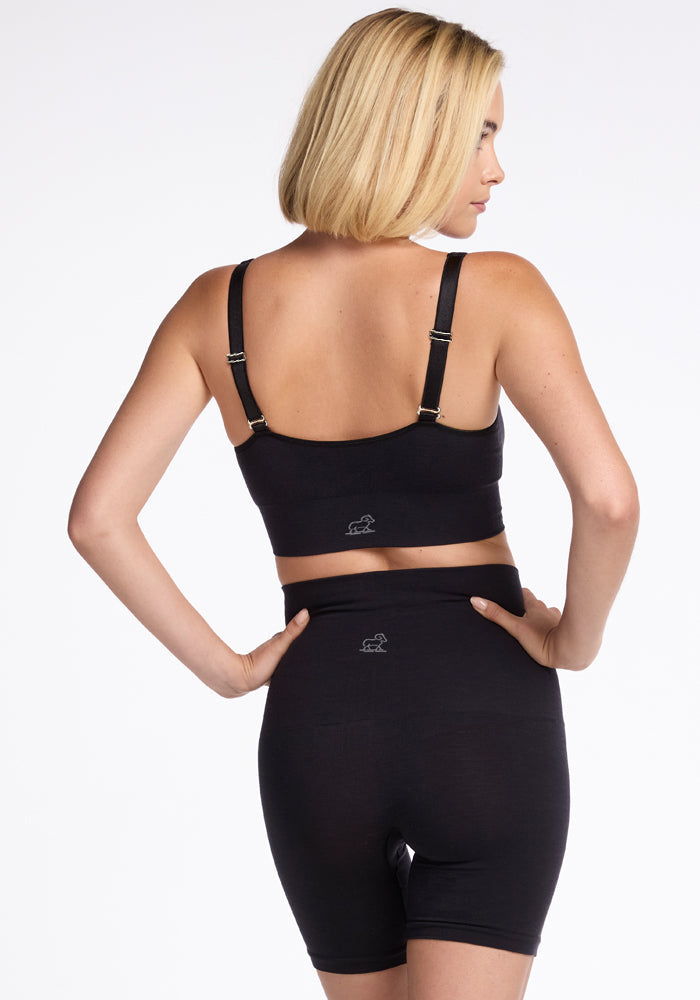 A woman with shoulder-length blonde hair stands with her back to the camera, showcasing a Woolx black bra and Marley Shapewear Shorts designed for breathable comfort. The plain white background highlights the sleek and stylish look of her outfit.