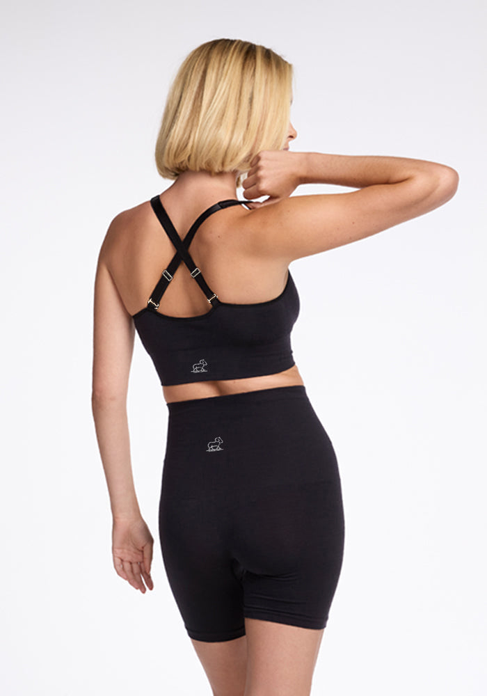 A person with short blonde hair is shown from the back wearing a black sports bra with cross-back straps and Woolx's Marley Shapewear Shorts. The high-waisted black shorts, adorned with small logo prints, feature breathable, lightweight merino wool undershorts. The background is plain white.