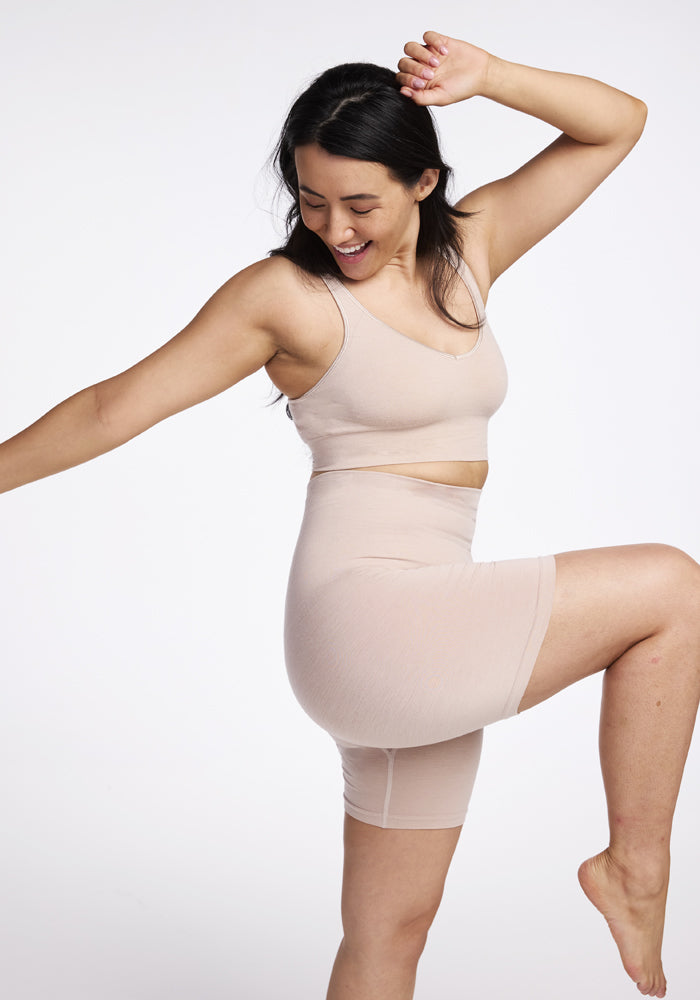 A person joyfully poses on one leg, dressed in a beige sports outfit from Woolx featuring the Hayden Bra in Sand paired with biker shorts. They smile with eyes closed and raise an arm in a carefree manner against a plain white background.