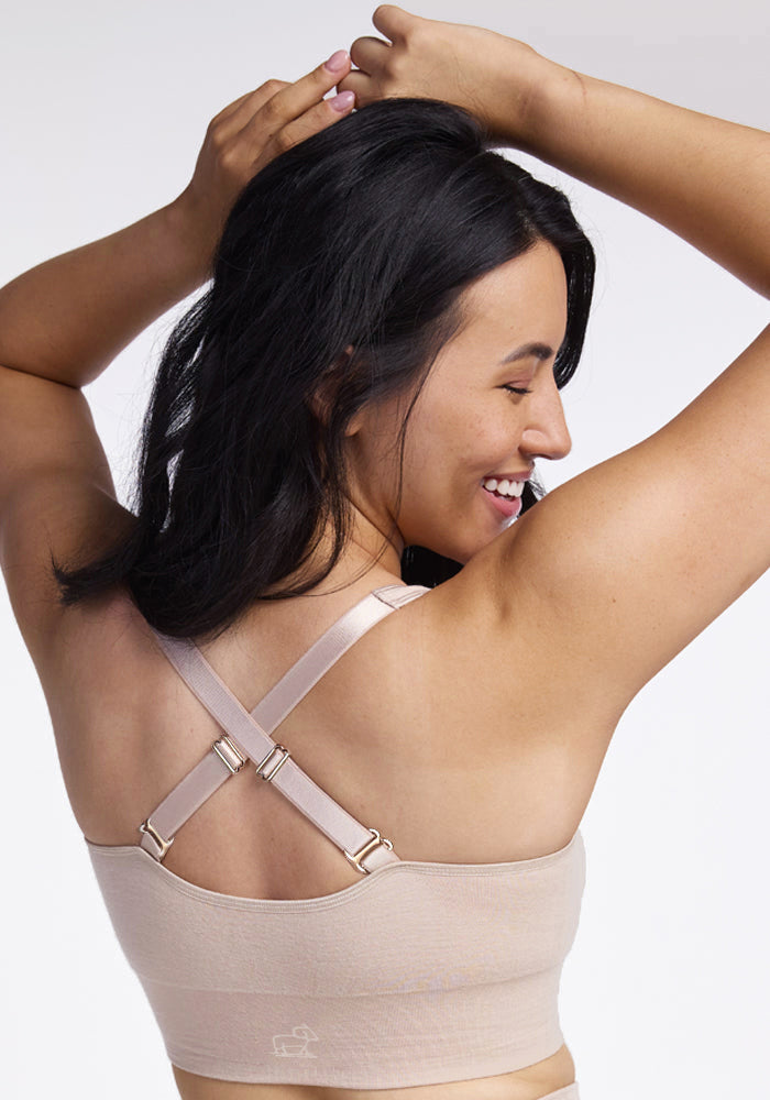 The woman with long dark hair is smiling and facing away from the camera, showcasing the Woolx Hayden Bra in Sand. Its seamless construction with crossed straps enhances comfort. Her raised arms highlight the quick-drying fabric against a plain light background.