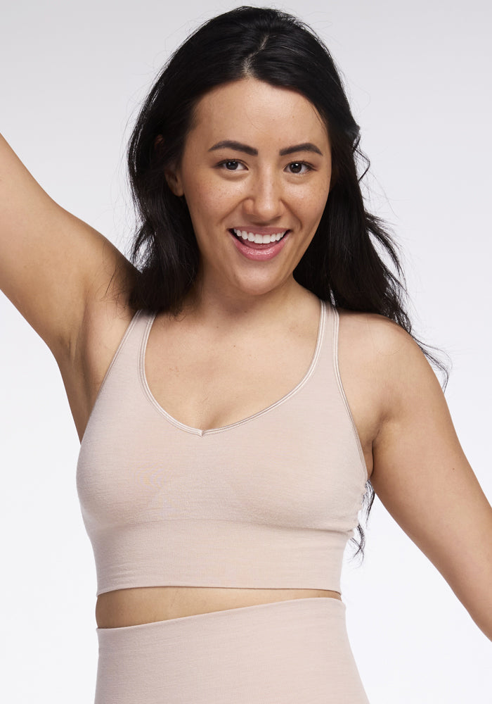 A person with long dark hair smiles and raises one arm, showcasing the seamless Hayden Bra - Sand by Woolx. They are posing against a plain white background, exuding confidence and elegance in their beige sportswear. 