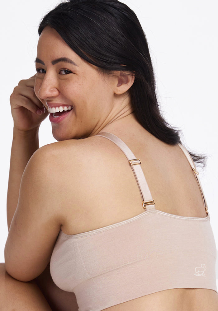 A woman with long dark hair is smiling and looking over her shoulder while wearing the cozy Woolx Hayden Bra, a lounge essential in beige. Seated and appearing happy and relaxed against a plain white background, she exudes effortless comfort.