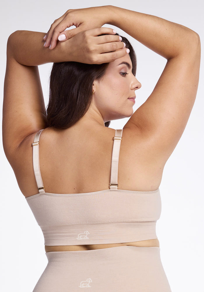 A woman is depicted from behind, dressed in a Sand Hayden Bra by Woolx with high-waisted bottoms characterized by seamless construction. Her arms are elevated, with her hands resting on the back of her head against a plain white backdrop.