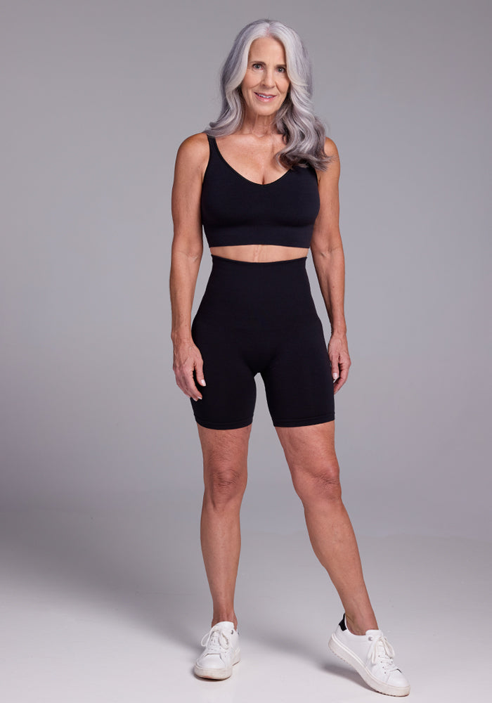 A woman with long gray hair, dressed in a black sports bra, Woolx Marley Shapewear Shorts, and white sneakers, stands against a plain gray background. She is smiling with one arm relaxed by her side and the other resting on her hip.