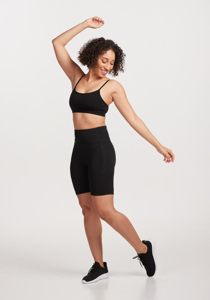 A person with curly hair is wearing a black sports bra and the Woolx MERINO FLEX™ Dani Bike Shorts - Black, striking a playful pose with arms raised. They have one leg bent and are smiling against a plain white background.