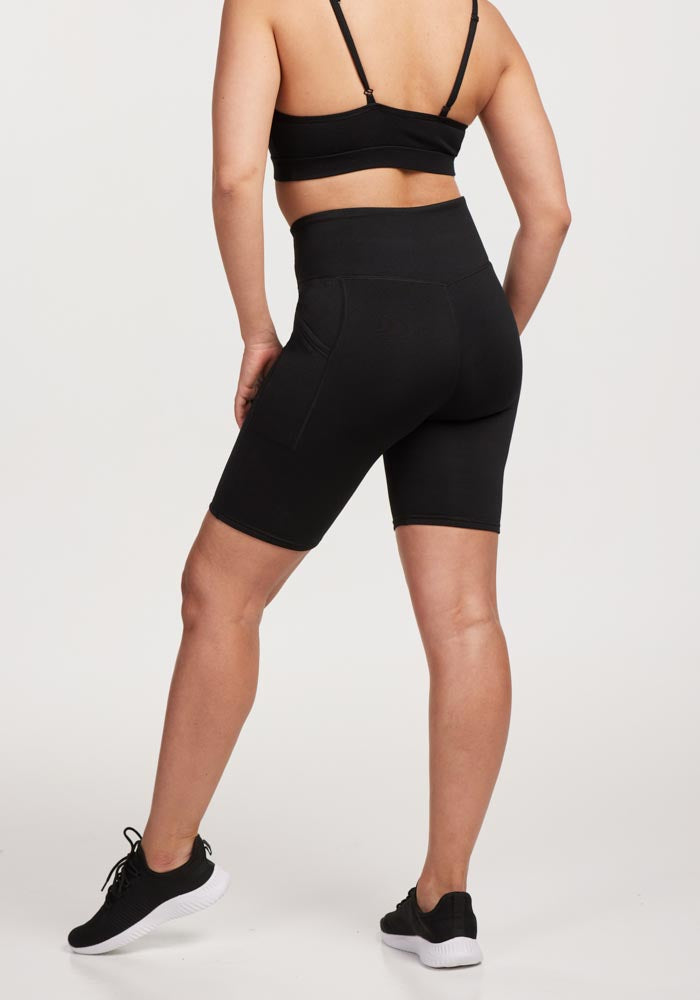 A person wearing the Woolx MERINO FLEX™ Dani Bike Shorts in black stands against a plain background. They face away, hands on hips, paired with sleek black athletic shoes—perfect for everyday wear with comfort in style.