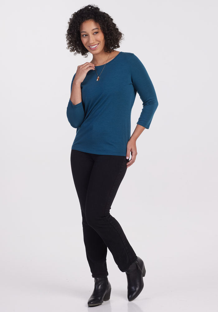 A person with curly hair is smiling and posing confidently in a Real Teal Jenny 3/4 Sleeve Crew Neck shirt by Woolx. They pair it stylishly with black pants and ankle boots against a plain white background.