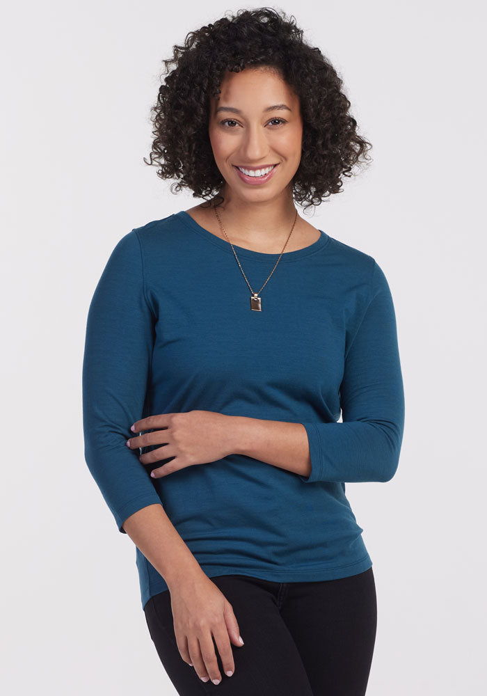 A person with curly hair smiles at the camera, adorned in a Real Teal Jenny 3/4 Sleeve Crew Neck by Woolx and a necklace. They stand against a plain white backdrop, effortlessly stylish in their lightweight top. 