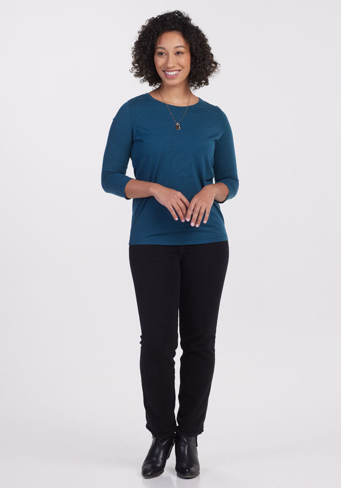 A person with curly hair is smiling, wearing the Jenny 3/4 Sleeve Crew Neck in Real Teal by Woolx, along with black pants and black ankle boots. They are standing against a plain white background, with their hands gently clasped in front.