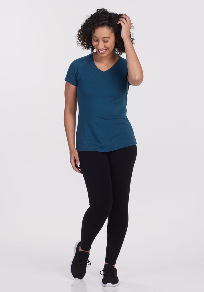 A person with curly hair is smiling and looking down while touching their hair. They are wearing the Woolx Mia V Neck shirt in Real Teal, crafted from merino wool, paired with black leggings and sneakers. This stylish outfit not only looks great but also stays odor-free – ideal for their next adventure.